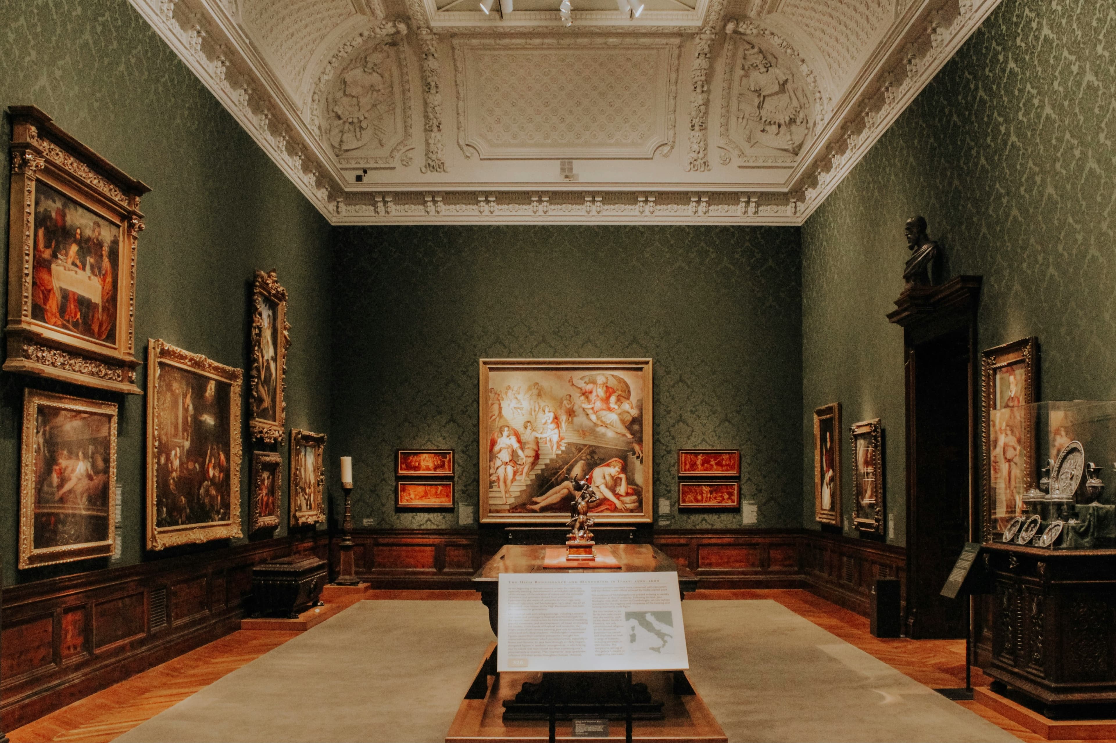 よその国の絵画館の展示室。緑のダマスク張りの壁に金彩の額の油彩が十数点、漆喰の格天井、天井からの自然光、大理石の胸像とガラスケース、中央に英語の解説パネル。